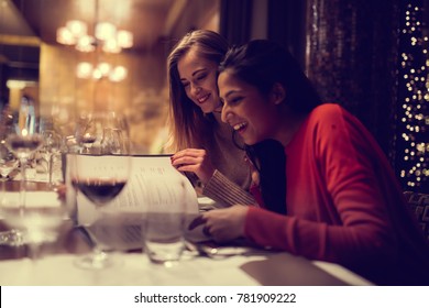 Two Adolescent Women Sitting In A Fancy Restourant, Going Through The Menue, While Drinking Some Red Wine, Talking, Gossiping And Unwrapping Their Christmass Presents And Looking At Their Phones. 