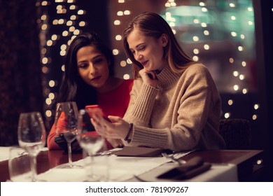 Two Adolescent Women Sitting In A Fancy Restourant, Going Through The Menue, While Drinking Some Red Wine, Talking, Gossiping And Unwrapping Their Christmass Presents And Looking At Their Phones. 