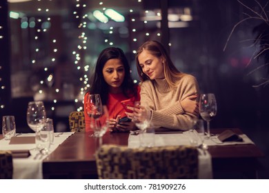 Two Adolescent Women Sitting In A Fancy Restourant, Going Through The Menue, While Drinking Some Red Wine, Talking, Gossiping And Unwrapping Their Christmass Presents And Looking At Their Phones. 