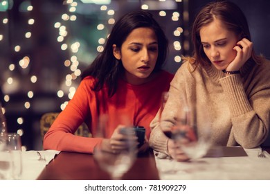 Two Adolescent Women Sitting In A Fancy Restourant, Going Through The Menue, While Drinking Some Red Wine, Talking, Gossiping And Unwrapping Their Christmass Presents And Looking At Their Phones. 