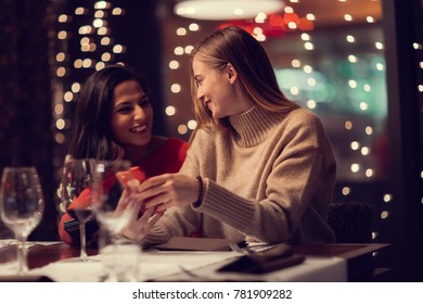 Two Adolescent Women Sitting In A Fancy Restourant, Going Through The Menue, While Drinking Some Red Wine, Talking, Gossiping And Unwrapping Their Christmass Presents And Looking At Their Phones. 