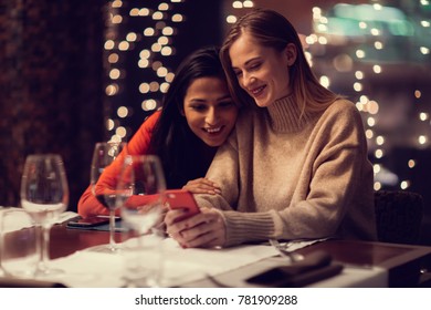 Two Adolescent Women Sitting In A Fancy Restourant, Going Through The Menue, While Drinking Some Red Wine, Talking, Gossiping And Unwrapping Their Christmass Presents And Looking At Their Phones. 