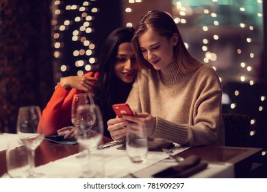 Two Adolescent Women Sitting In A Fancy Restourant, Going Through The Menue, While Drinking Some Red Wine, Talking, Gossiping And Unwrapping Their Christmass Presents And Looking At Their Phones. 