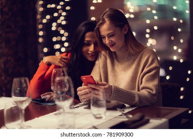 Two Adolescent Women Sitting In A Fancy Restourant, Going Through The Menue, While Drinking Some Red Wine, Talking, Gossiping And Unwrapping Their Christmass Presents And Looking At Their Phones. 