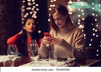 Two Adolescent Women Sitting In A Fancy Restourant, Going Through The Menue, While Drinking Some Red Wine, Talking, Gossiping And Unwrapping Their Christmass Presents And Looking At Their Phones. 