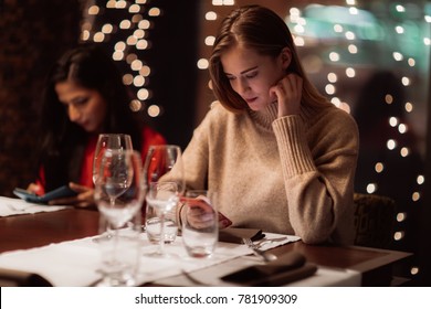 Two Adolescent Women Sitting In A Fancy Restourant, Going Through The Menue, While Drinking Some Red Wine, Talking, Gossiping And Unwrapping Their Christmass Presents And Looking At Their Phones. 