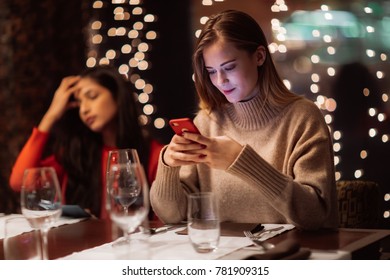 Two Adolescent Women Sitting In A Fancy Restourant, Going Through The Menue, While Drinking Some Red Wine, Talking, Gossiping And Unwrapping Their Christmass Presents And Looking At Their Phones. 