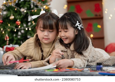 Two adorable and happy young Asian girls are enjoying painting their Christmas cards with pencil colors, having fun, and celebrating Christmas at home together.