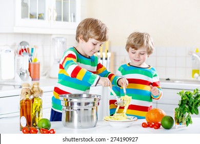 Two Adorable Little Boys Cooking And Eating Meal With Spaghetti And Fresh Vegetables In Domestic Kitchen, Indoors. Sibling Children In Colorful Shirts.