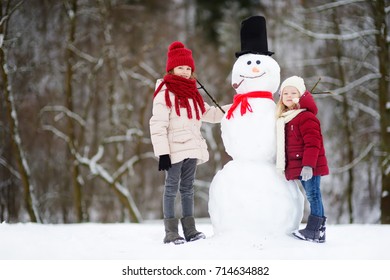 Two Adorable Little Girls Building A Snowman Together In Beautiful Winter Park. Cute Sisters Playing In A Snow. Winter Activities For Kids.