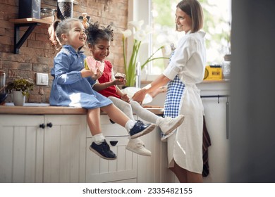 Two adorable little girls sitting on kitchen counter eating watermelon and muffin together, with their mother by their side washing dishes in a modern kitchen. Home, family, lifestyle concept. - Powered by Shutterstock