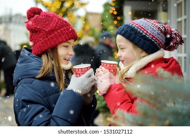 Two Adorable Sisters Drinking Hot Chocolate On Traditional Christmas Fair In Riga, Latvia. Children Enjoying Sweets, Candies And Gingerbread On Xmas Market. Winter Time With Family And Kids.