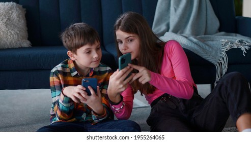Two adorable teenage kids using smartphones staying at home. Brother and elder sister playing mobile phone games online together. Living room. Apartment.