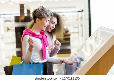 Two adult caucasian girlfriends use information kiosk at shopping center. Happy women with shopping bags pointing finger at the info desk. Consumerism, shopping, lifestyle concept - Powered by Shutterstock