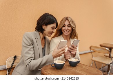 Two adult caucasian women expressing fun use mobile phones. Ladies in casual clothes sit at table drinking coffee. People emotions and spring holiday concept. - Powered by Shutterstock