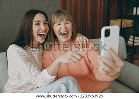 Two adult laughing women mature mom young kid wearing casual clothes hold use mobile cell phone sit on gray sofa couch stay at home flat rest relax spend free spare time in living room. Family concept