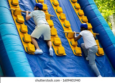 Two Adult Men Having Funny Competition In Climbing On Inflatable Castle With Wooden Sticks In Outdoor Amusement Playground

