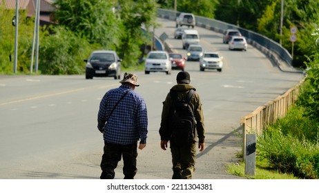 Two Adult Men Are Walking Along The Road On A Sunny Day, Cars Are Driving Towards The Meeting, View From The Back.