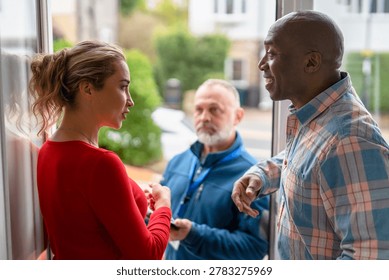Two adults are having a conversation while a man stands nearby looking at them. The setting is a residential area in broad daylight.