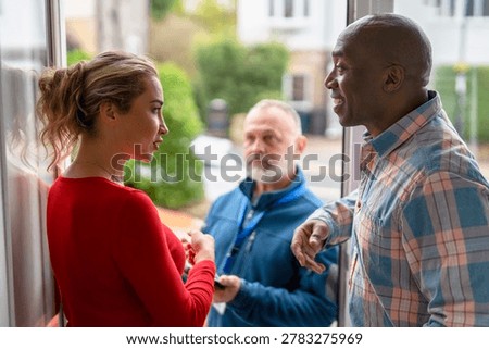 Two adults are having a conversation while a man stands nearby looking at them. The setting is a residential area in broad daylight.