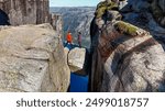 Two adventurers carefully walk across a rocky ledge, with Kjeragbolten balancing dramatically between cliffs above a blue fjord. a couple of men and women at Kjerag Kjeragbolten Lysefjord Norway