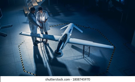 Two Aerospace Engineers Work On Unmanned Aerial Vehicle / Drone Prototype. Aviation Scientists In White Coats Talking, Using Tablet Computer. Industrial Laboratory With Surveillance Military Aircraft