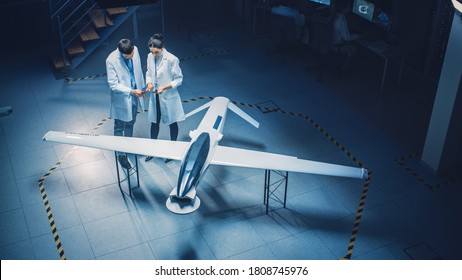 Two Aerospace Engineers Work On Unmanned Aerial Vehicle Drone Prototype. Aviation Scientists in White Coats Talking, Using Tablet Computer. Industrial Laboratory with Surveillance or Military Aircraft