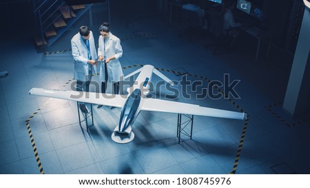 Two Aerospace Engineers Work On Unmanned Aerial Vehicle Drone Prototype. Aviation Scientists in White Coats Talking, Using Tablet Computer. Industrial Laboratory with Surveillance or Military Aircraft
