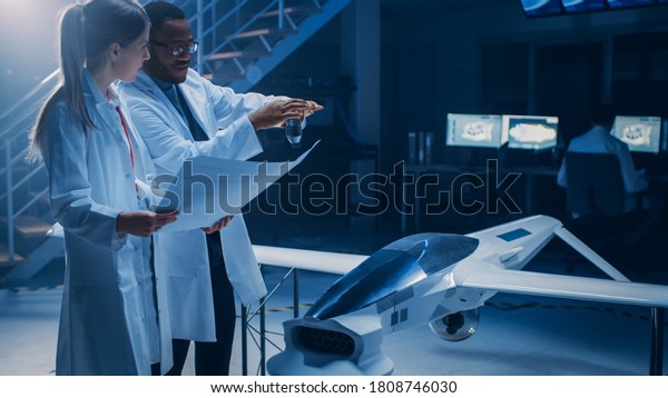 Two Aerospace Engineers Work On Unmanned Aerial\
Vehicle / Drone Prototype. Aviation Scientists Talking, using\
Blueprints. Industrial Laboratory with Commercial Aerial\
Surveillance Aircraft