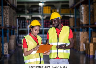 Two African America Engineer Wearing Safety Helmet And Vest Standing In The Automotive Warehouse With Blur Background. Woman Holding Clipboard And Discuss With Man. Distribution And Inventory Concept