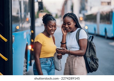 Two African american female friends waiting for a bus while at a bus stop and using a smartphone together. Riding, sightseeing, traveling to work, city tour, togetherness. Copy space. - Powered by Shutterstock