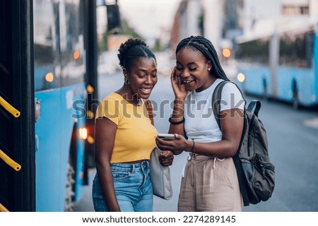 Two African american female friends waiting for a bus while at a bus stop and using a smartphone together. Riding, sightseeing, traveling to work, city tour, togetherness. Copy space.