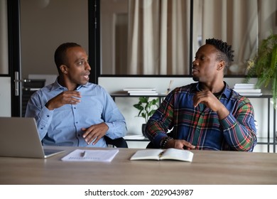 Two African American male employees sit at desk in office cooperate work together on computer. Mixed race men colleagues brainstorm negotiate on laptop, engaged in teamwork at workplace.