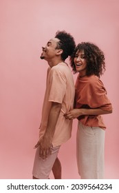 two African American men stand sideways on gently pink studio background. cute lady in carrot-colored T-shirt tickles her bearded friend in peach-colored T-shirt.