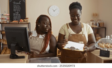 Two african american women bakers working together in a bakery shop, one using a computer while the other holds a clipboard, surrounded by pastries, bread, and a gluten-free sign. - Powered by Shutterstock