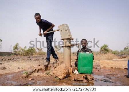 Two African boys busy filling water containers at a remote village pump; water scarcity in developing countries concept