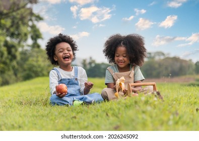 Two African Dark Skinned Children Boy And Girl Sitting On Field Grass And Eating Fruits Together In Parks And Outdoors. Happy Family African Kids Boy And Girl Eat Apple And Banana Sit Picnic On Meadow