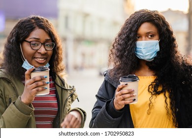 Two African Female Friends Having Drinks Outdoors During Travel In European City. Happy Nigerian Women Tourists In Protective Mask Laughing Cheerful. Girls Holding Paper Cups Of Coffee.