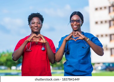 Two African Health Professionals In Scrubs Showing Love Sign With Hands At Hospital Premises - Black Medical Students On University Campus