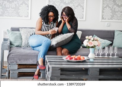 Two african woman friends wear on eyeglasses sitting at couch indoor white room and looking with surprised faces at mobile phone.