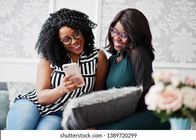 Two African Woman Friends Wear On Eyeglasses Sitting At Couch Indoor White Room And Looking At Mobile Phone.