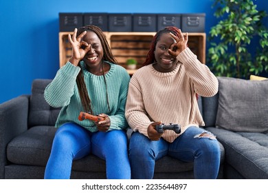 Two african woman playing video game holding controller smiling happy doing ok sign with hand on eye looking through fingers  - Shutterstock ID 2235644897