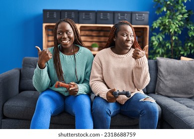 Two african woman playing video game holding controller pointing thumb up to the side smiling happy with open mouth  - Shutterstock ID 2318688759