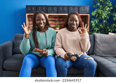 Two african woman playing video game holding controller doing ok sign with fingers, smiling friendly gesturing excellent symbol  - Shutterstock ID 2334641917