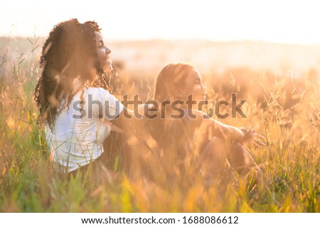two afro american girls sitting on the field looking at the sunset with the golden sunlight iluminating the scene with their right side facing the camera. Imagine de stoc © 