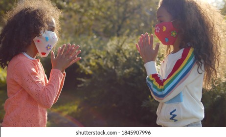 Two Afro Girls In Safety Mask Playing Hand Clapping Game Outdoors. Side View Of African Sister Wearing Protective Mask Having Fun Together Outside On Summer Day