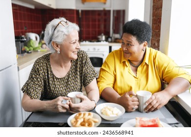 Two aged ladies of diverse ethnicity sitting at kitchen table drinking coffee, sharing gossips and rumors, discussing news, looking at each other with laughing faces, enjoying spending time together - Powered by Shutterstock