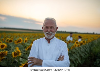 Two Agronomists Standing In Sunflower Field Talking In Background. Senior Biologist Smiling Looking At Camera With Arms Crossed.