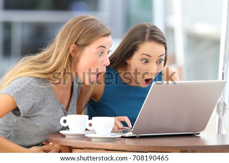 Two amazed friends watching scandalous content in a laptop sitting in a bar