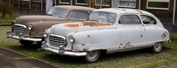 Two American Cars From The 1950s, Old And Rusty Without Paint, Brown And White, Windows Are Partly Broken With Holes, Front Right Photographed, On The Day Without People